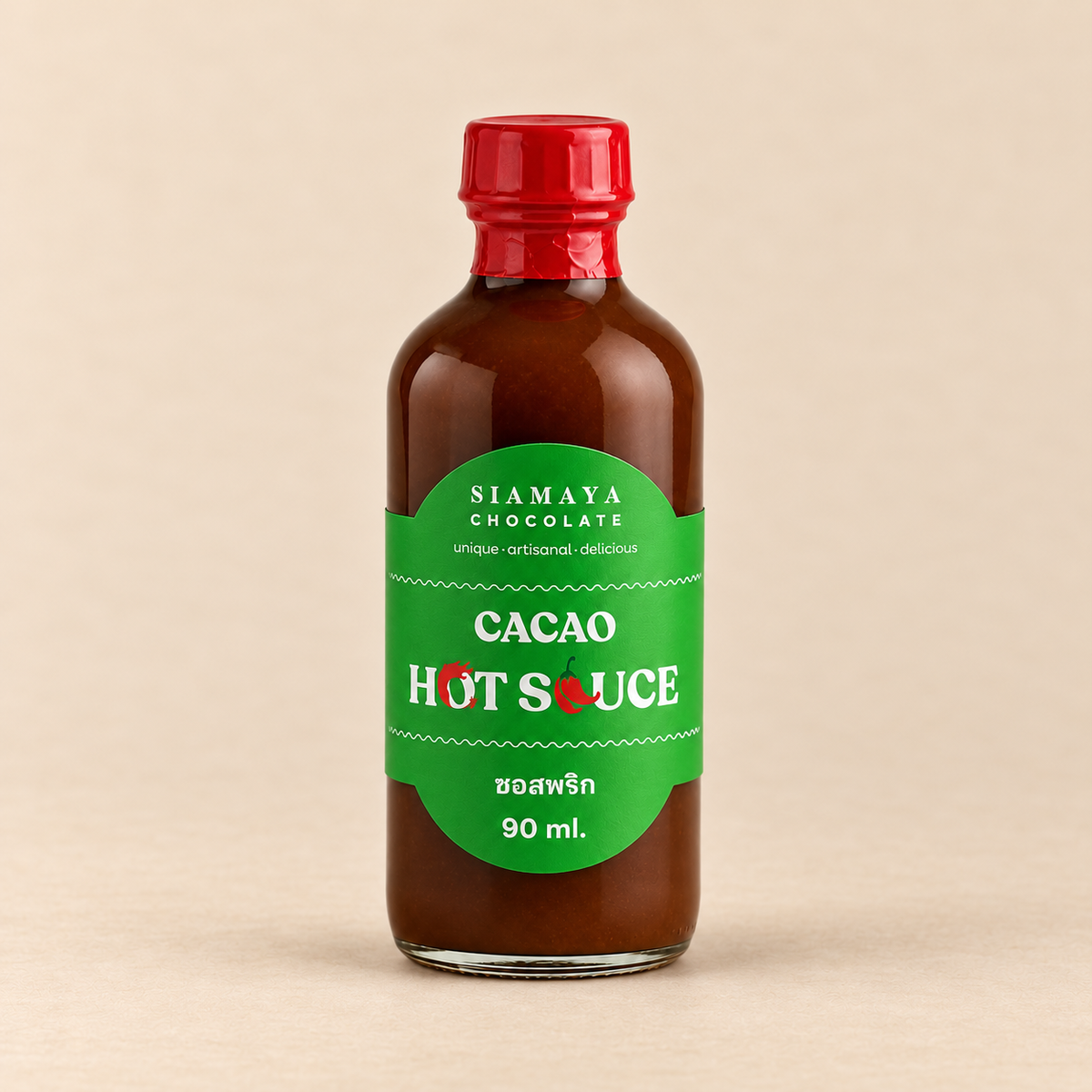 Siamaya Cacao Hot Sauce, 90 ml bottle with green label and red cap, on a warm sand studio background.