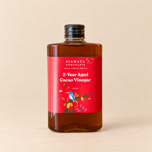 Siamaya 2-Year Aged Cacao Vinegar, 200 ml bottle with red illustrated label, on a warm sand studio background.
