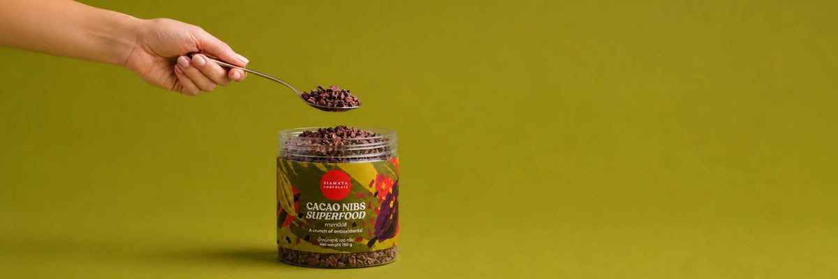 A hand lifting a spoonful of cacao nibs from a jar of Siamaya Cacao Nibs Superfood