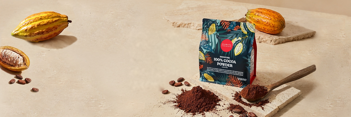 A pouch of Siamaya Signature 100% Cocoa Powder on pale stone, with cocoa powder, a wooden spoon and cacao pods