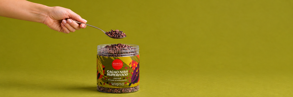 A hand holds a spoonful of cacao nibs above an open Siamaya Cacao Nibs Superfood 150 g jar on an olive green background.