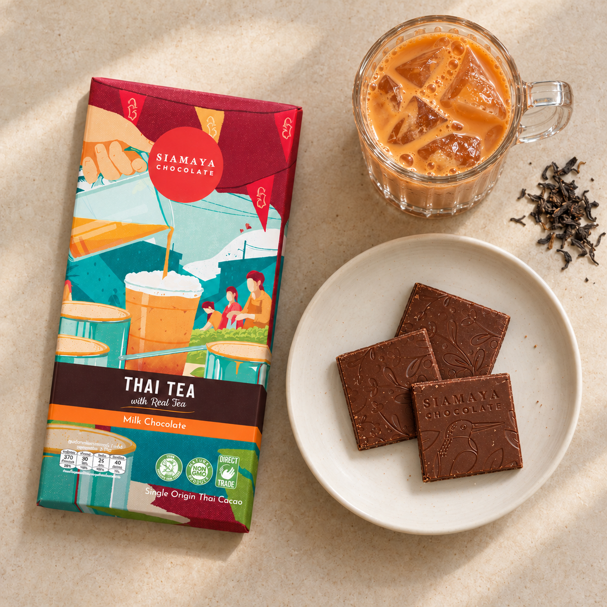 Siamaya Thai Tea Milk Chocolate wrapper with three chocolate squares on a plate, iced Thai tea in a faceted handled glass, and loose tea on a light café tabletop.